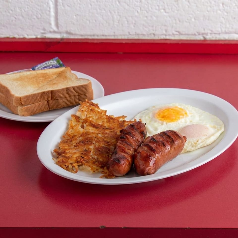 Breakfast plate with two sausages, fried egg, and hash browns, accompanied by toasted bread on a red diner table.