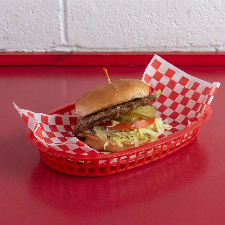 Double cheeseburger with lettuce, tomato, pickles, and onions served in a red basket lined with checkered paper.