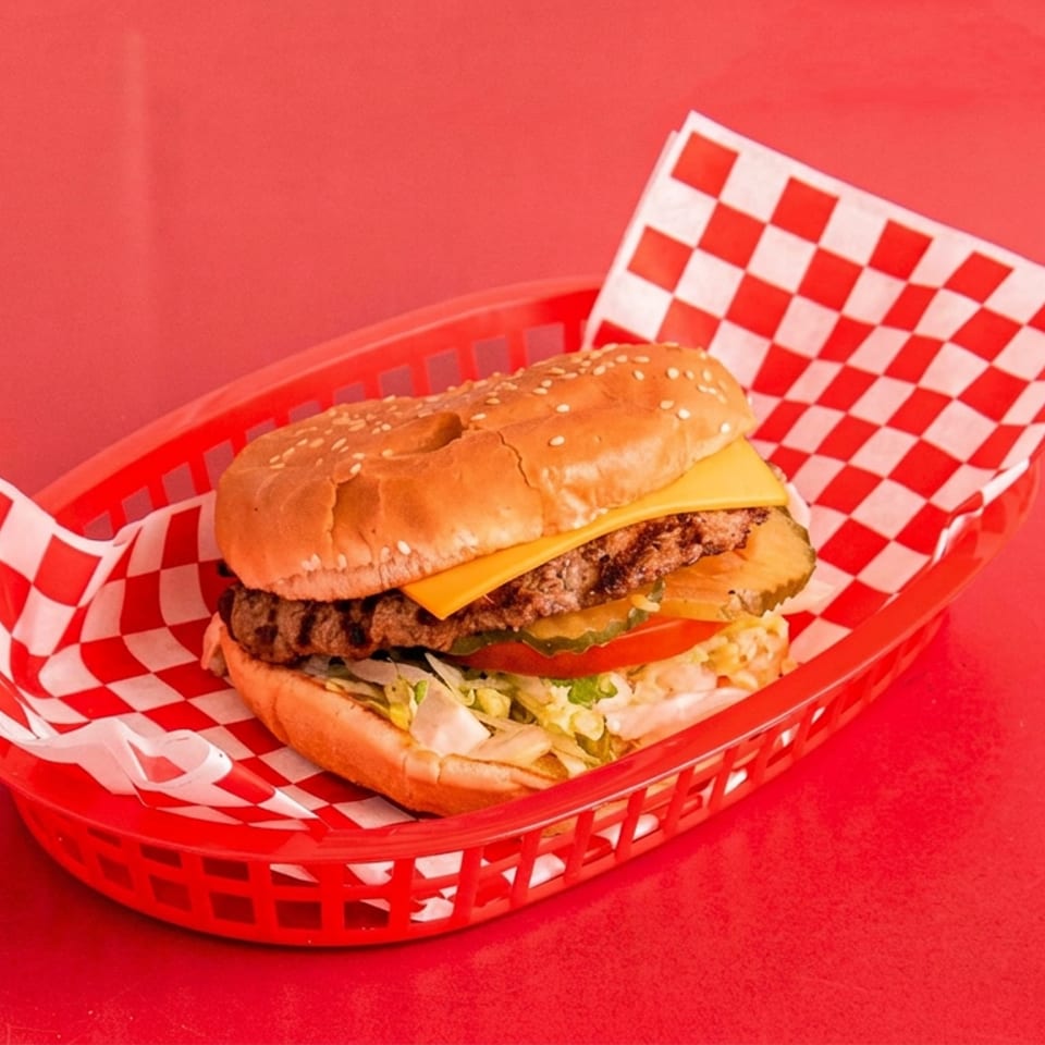 Cheeseburger with lettuce, tomato, pickles, and onions in a red basket lined with checkered paper.